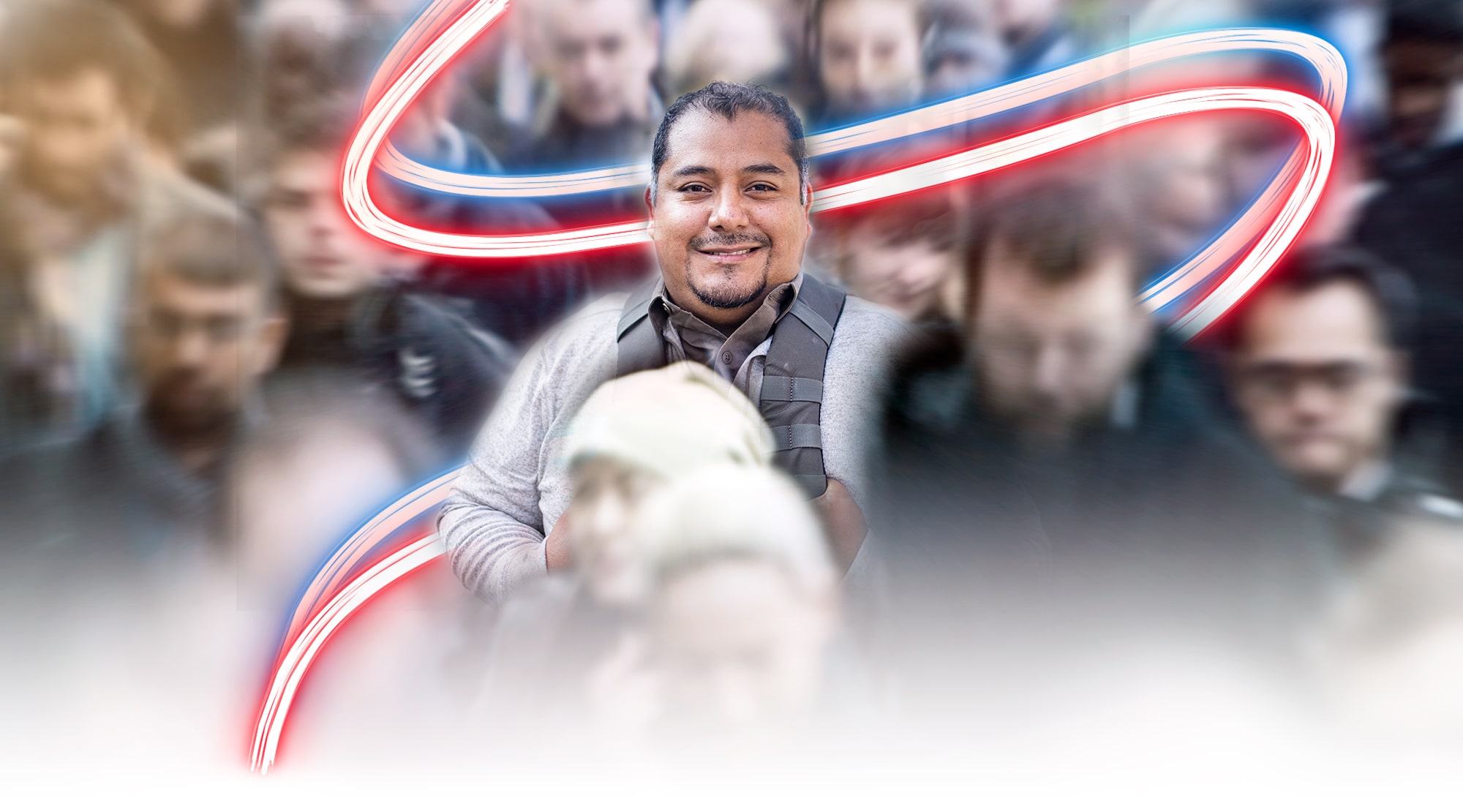 A Veteran in a grey cardigan over a similarly colored shirt stands out from the center of a crowd of people. He wears a backpack and is slightly smiling into the camera. The crowd he stands out from is intentionally blurred for artistic effect. A neon stripe of red and blue meanders down the image behind the man.